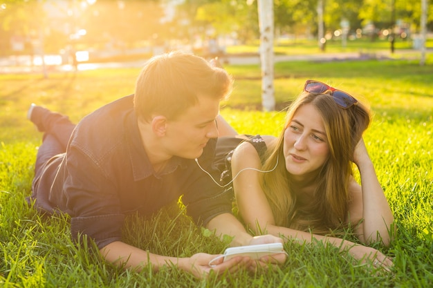 Sunny portrait of sweet young couple lying relaxing on the grass together listens to music in earphones on smartphone, summer day.