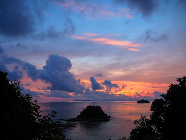 Sunrise sea view with small island and colorful sky through tree leaves silhouette