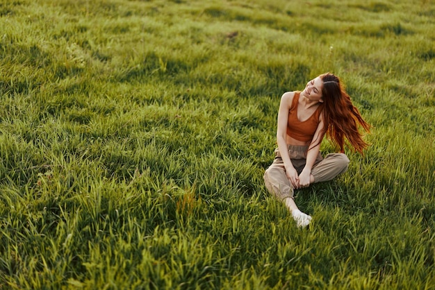 The sunset light of a summer evening illuminates a beautiful young redheaded woman sitting on the grass in green pants and an orange top and smiling at the sun