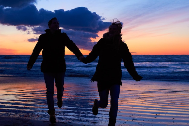 Sunset silhouette and couple running at beach on romantic date vacation or holiday in nature Man and woman holding hands with love and care on travel adventure or trip with clouds sky and ocean