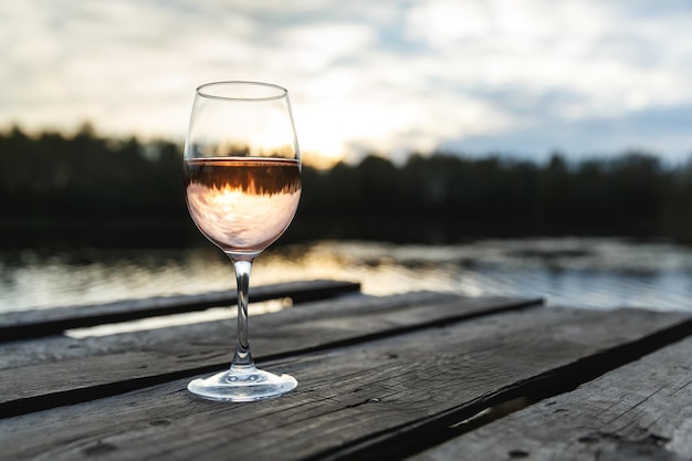 Sunset view of a glass of orange wine on a wooden pier on a lake. Wine drinking on a beautiful location