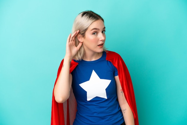 Super Hero caucasian woman isolated on blue background listening to something by putting hand on the ear