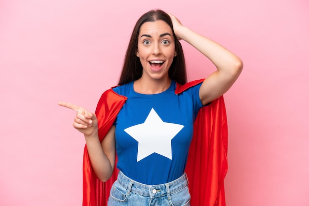 Super Hero Caucasian woman isolated on pink background surprised and pointing finger to the side