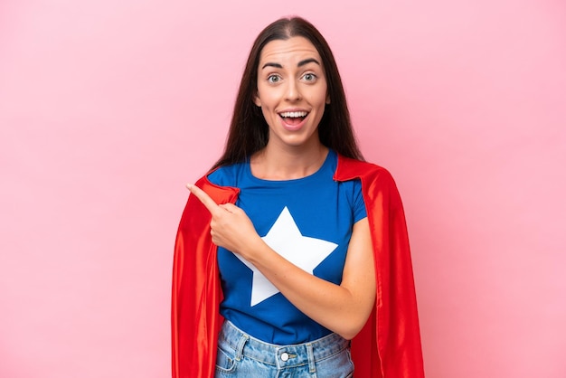 Super Hero Caucasian woman isolated on pink background surprised and pointing side