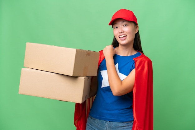 Super Hero delivery Asian woman holding boxes over isolated background celebrating a victory