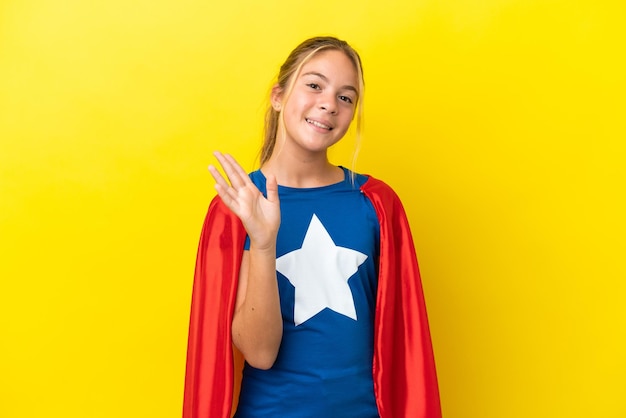 Super Hero little girl isolated on yellow background saluting with hand with happy expression