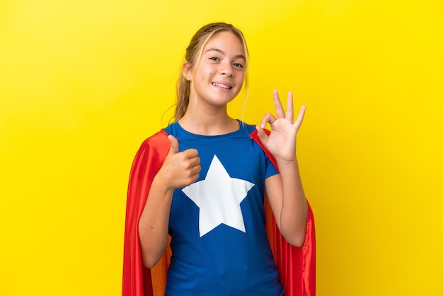 Super Hero little girl isolated on yellow background showing ok sign and thumb up gesture