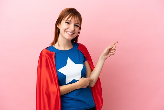 Super Hero redhead girl isolated on pink background pointing finger to the side