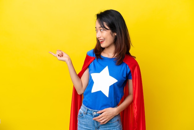 Super hero Vietnamese woman isolated  on yellow background pointing finger to the side and presenting a product