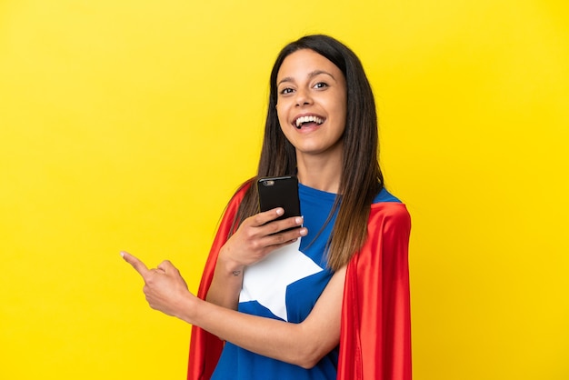 Super Hero woman isolated on yellow background using mobile phone and pointing back