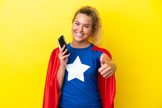 Super Hero woman isolated on yellow background using mobile phone while doing thumbs up