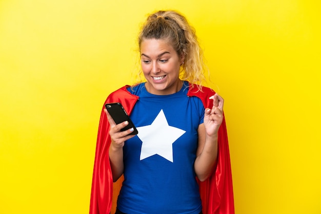 Super Hero woman isolated on yellow background using mobile phone with fingers crossing