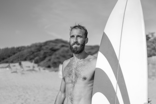 Surfer holding his surfboard on the beach Hipster man standing on the beach and waiting big waves for surfing Fit bearded man training with surfboard to sea Lifestyle and freedom concept