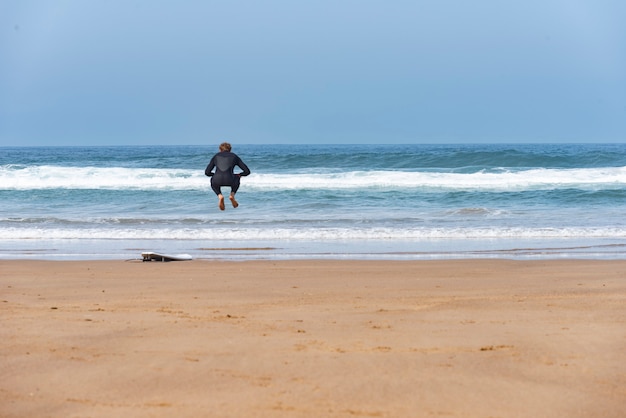 Surfer jumping on the beach next to his board with the sea in the background