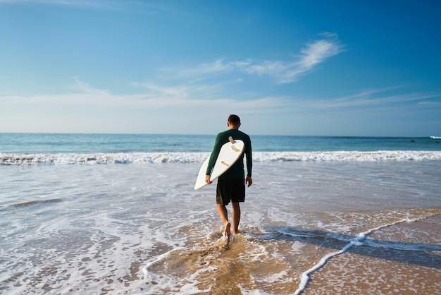 Surfer man walks into ocean with board at sunrise Adventurous male heads to lineup for surf session