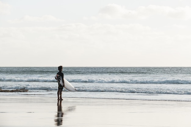 A surfer watching the waves standing on the beach shore at sunset