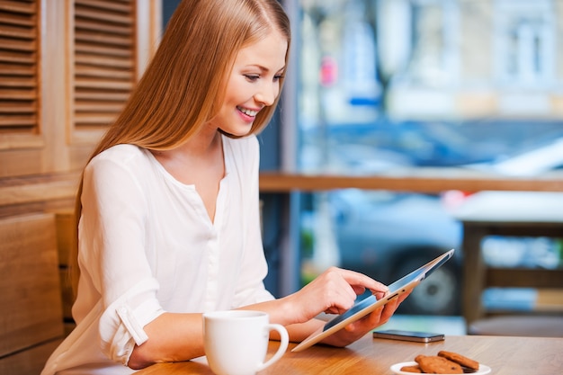 Surfing the net in cafe. Side view of beautiful young woman using digital tablet and smiling while enjoying coffee in cafe