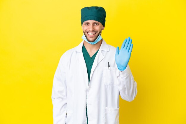 Surgeon blonde man in green uniform isolated on yellow background saluting with hand with happy expression