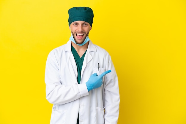 Surgeon blonde man in green uniform isolated on yellow background surprised and pointing side