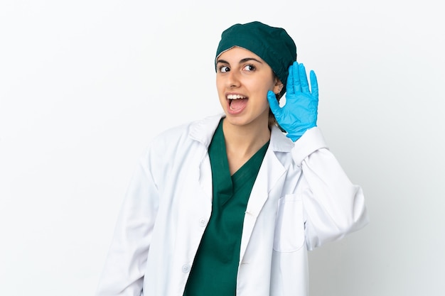 Surgeon caucasian woman isolated on white background listening to something by putting hand on the ear