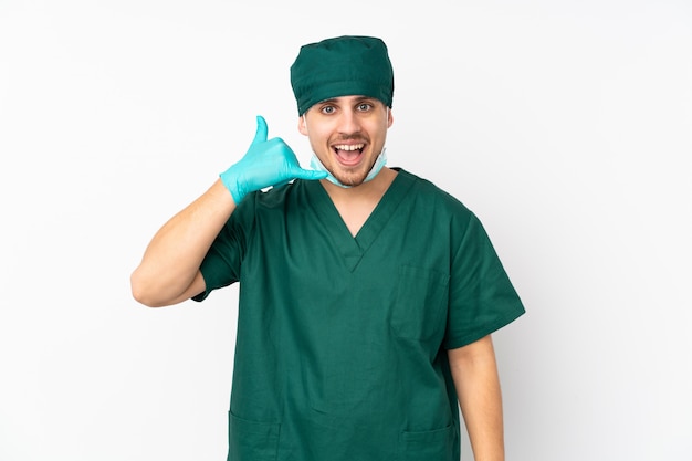 Surgeon in green uniform on isolated white wall making phone gesture.