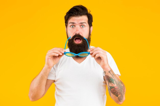 Surprise party Surprised hipster hold glasses yellow background Bearded man show surprise on unshaven face Keeping mouth opened of surprise Surprise emotions