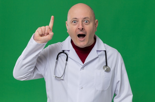 Surprised adult man in doctor uniform with stethoscope pointing up 