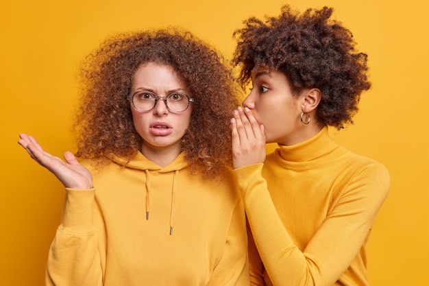 Photo surprised afro american woman whispers secret to friend tells secretly something on ear share gossips dressed casually isolated over yellow wall. puzzled curly european female hears rumor