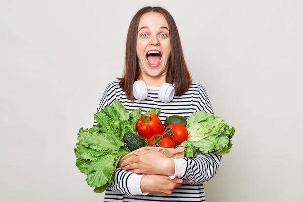 Surprised amazed woman with brown hair wearing striped shirt standing isolated over gray background holding vegetables screaming with excitement