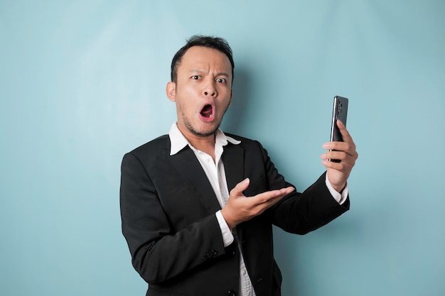 Surprised Asian businessman wearing black suit holding his smartphone isolated by blue background