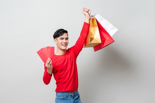 Surprised Asian man raising hands showing shopping bags and red envelope Ang Pow in isolated light gray studio background for Chinese new year sale concept