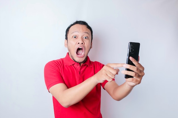 Surprised Asian man wearing red tshirt pointing at his smartphone isolated by white background