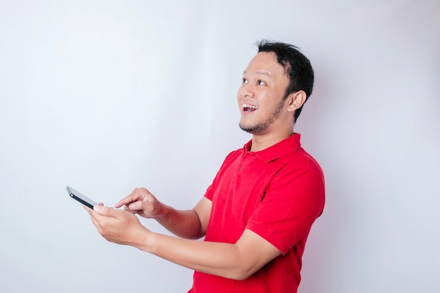 Surprised Asian man wearing red tshirt pointing at his smartphone isolated by white background