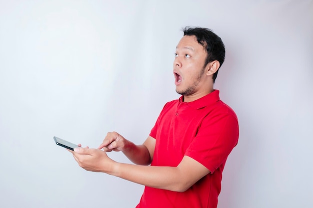 Surprised Asian man wearing red tshirt pointing at his smartphone isolated by white background