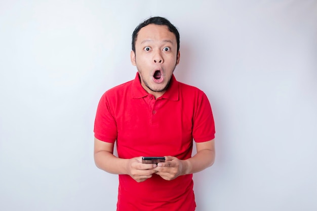 Surprised Asian man wearing red tshirt pointing at his smartphone isolated by white background