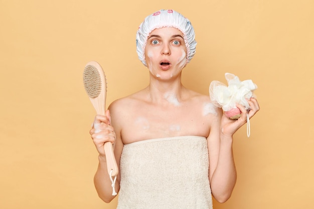 Photo surprised astonished woman wearing shower cap standing isolated over beige background holding washcloth and brush for dry massage