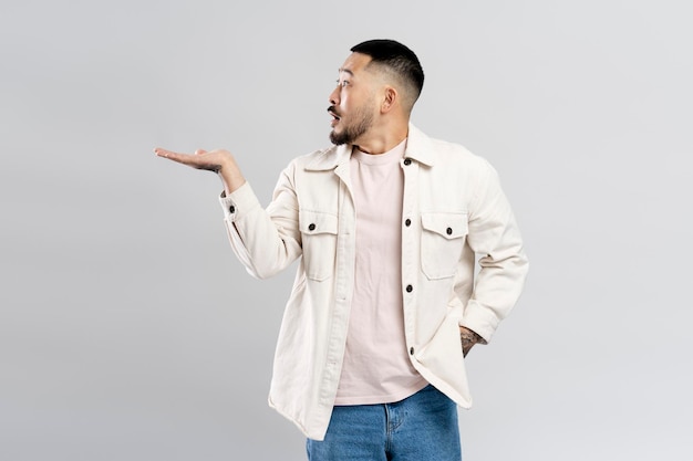 Surprised attractive asian man pointing with hand at copy space isolated on gray background