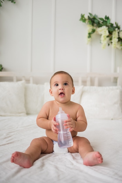 Surprised baby boy in a diaper sits on the bed and holds a bottle of water