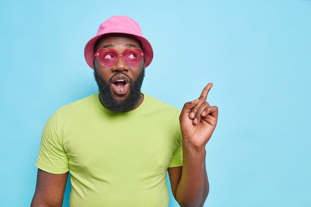 Surprised bearded man points at upper right corner shows something breathtaking attracts your attention to advertisement wears casual t shirt and panama isolated over blue wall