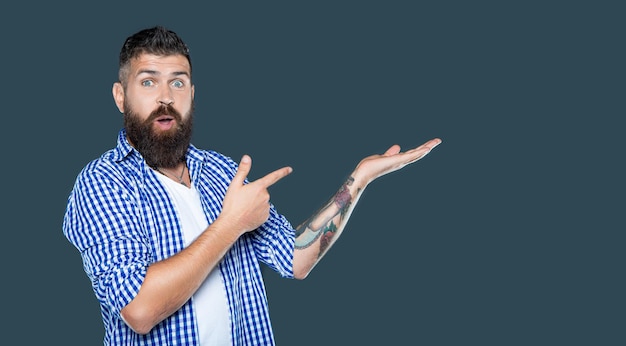 Surprised bearded man wear checkered shirt presenting product on grey background with copy space