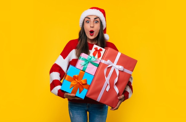 Surprised beautiful excited smiling woman with many cool Christmas gift boxes in hands while she is having fun and posing on yellow background