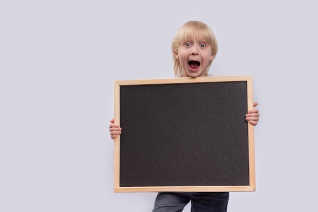 Surprised blond boy holding blackboard in hands. Copy space. Template. Mockup