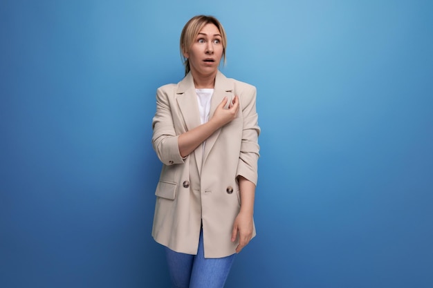 Surprised blond young woman in jacket pointing finger to side on studio background