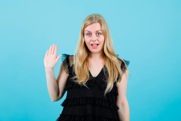 Surprised blonde girl is looking at camera by hsowing hi gesture on blue background