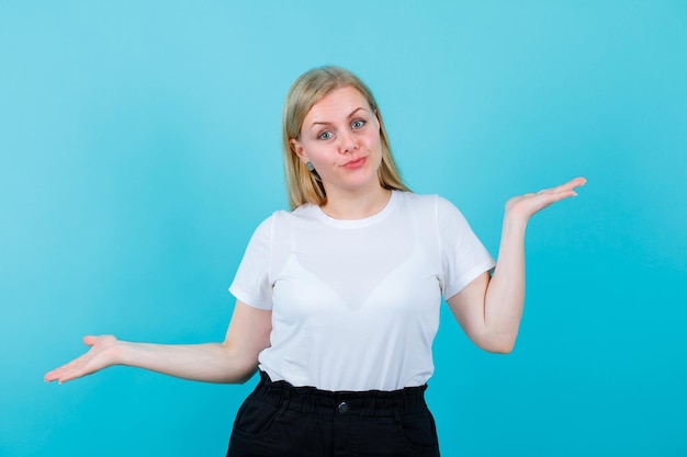 Surprised blonde girl is looking at camera by opening wide her arms on blue background