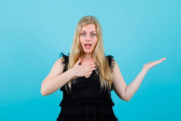 Surprised blonde girl is pointing right with hand on blue background