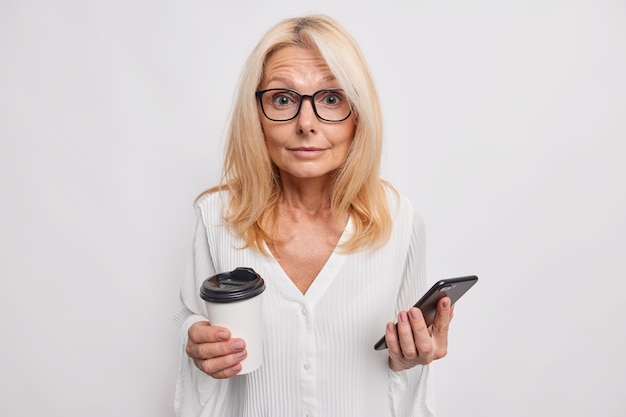 Surprised blonde middle aged woman messaging on smartphone sends sms drinks takeaway coffee wears glasses and blouse isolated over white wall distracted from chatting online enjoys leisure time