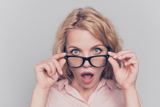 Surprised blonde touching glasses wide open eyes and mouth isolated on gray