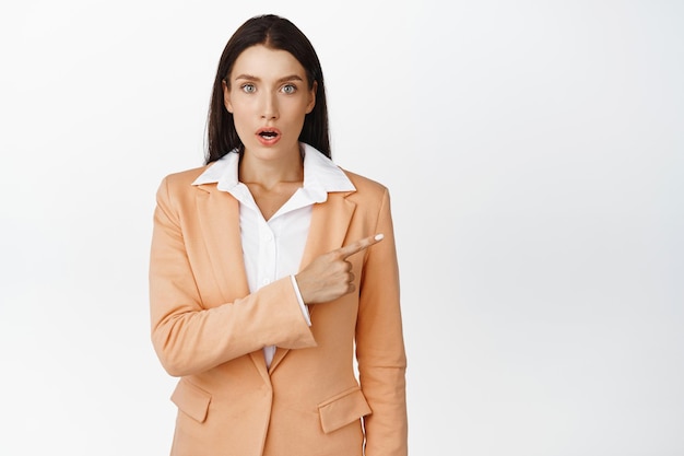 Surprised brunette businesswoman pointing right looking amazed at camera gasping and staring in awe standing in suit over white background