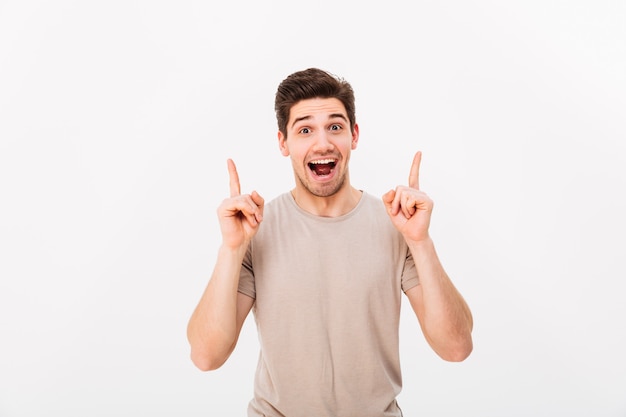 Surprised brunette man 30s wearing beige t-shirt gesturing fingers upward on copyspace with excitement on face, isolated over white wall